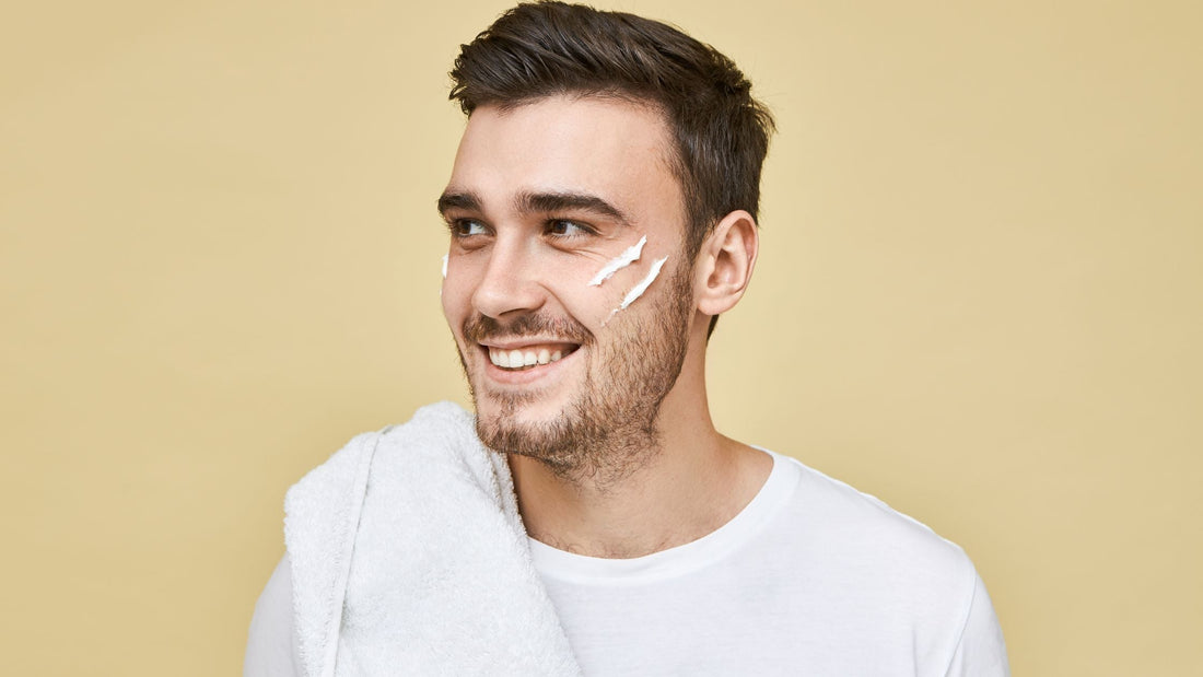 Man with stubble applying sunscreen for men's face with two finger stripes on cheek, holding white towel, smiling against beige background.