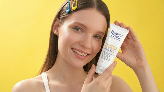 Smiling woman holding Heaven Roots Nia Defense SPF 50 PA++++ tube against a bright yellow background, showcasing the BEST Non-Comedogenic SUNSCREEN for lightweight, no white cast daily sun protection.